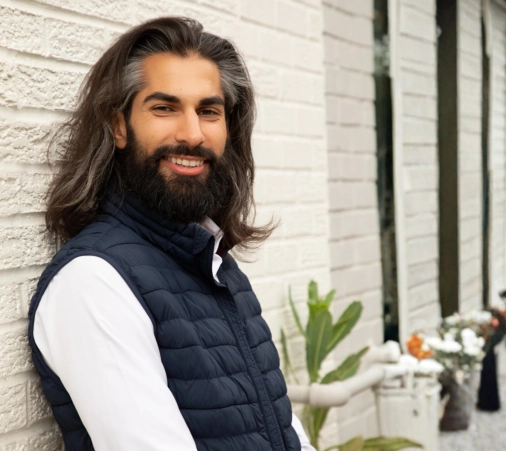 A smiling man with long hair and a beard leans against a white brick wall, wearing a dark blue vest and white shirt. The mood is relaxed and friendly-Homebuyer-Wallet-by-Kameron-Kang