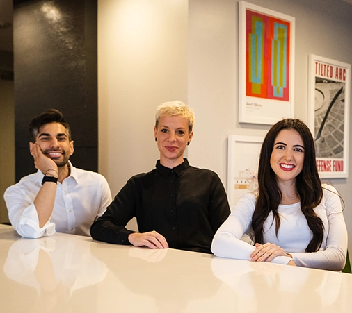 About Us 1 Three people, two women and a man, smiling and seated at a white table in a modern office with colorful artwork on the walls, convey a friendly, professional atmosphere
