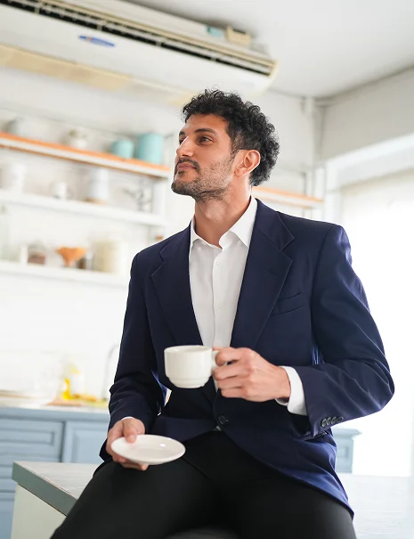 A smiling man with long hair and a beard leans against a white brick wall, wearing a dark blue vest and white shirt. The mood is relaxed and friendly-Homebuyer-Wallet-by-Kameron-Kang